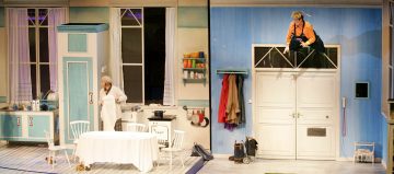 Keittiötä ja ruokailutilaa muistuttava värikäs lavaste, jossa yksi näyttelijä seisoo pöydän vieressä ja toinen oranssiin pukeutunut näyttelijä kiipeää oven yläpuolella olevasta yläikkunasta.