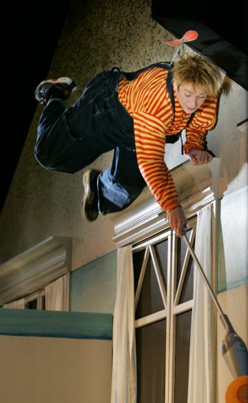 Haalareihin ja punakeltaiseen raidalliseen paitaan pukeutunut henkilö näyttää leijuvan vaakatasossa ikkunan yläpuolella ja kurottautuvan alaspäin pitkävartisen työkalun kanssa, mikä viittaa oudon tai teatterimaisen kohtauksen syntyyn.