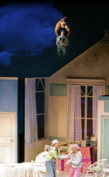 Kaksi Peter Paniksi ja toiseksi hahmoksi pukeutunutta ihmistä lentää yöllisen makuuhuoneen lavastuksen yläpuolella, kun taas kaksi värikkäisiin pukuihin pukeutunutta näyttelijää seisoo alhaalla ja katsoo yllättyneenä ylöspäin. Lavastuksessa on siniset seinät, ikkunat ja makuuhuoneen huonekalut.
