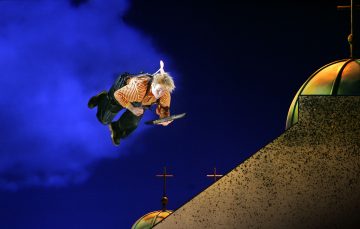 Raitapaitainen ja haalarimallinen henkilö lentää yöllä, kirja kädessään, potkuri selässään, risteillä koristeltujen kupolien yläpuolella syvänsinistä taivasta vasten.