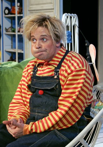 Koomisesti pukeutunut henkilö, jolla on sotkuiset vaaleat hiukset, punakeltainen raitapaita ja farkkuhaalari, istuu paisuneet posket ja katsoo sivulle humoristinen ilme leikkisässä, värikkäässä huoneessa.