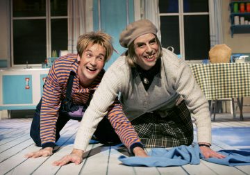 Kaksi värikkäisiin, leikkisiin pukuihin pukeutunutta ihmistä ryömii kirkkaan, sarjakuvamaisen keittiön lattialla hymyillen ja nauraen. Vasemmalla olevalla henkilöllä on raidallinen haalari, ja toisella on baskeri ja ruudullinen hame.