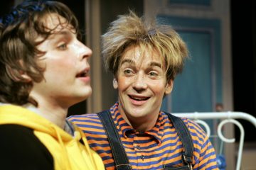 Kaksi ihmistä sisätiloissa, toinen etualalla keltainen huppari yllään, hieman epätarkka, ja toinen taustalla, jolla on sotkuinen vaalea tukka, yllään raidallinen oranssin ja sinisen värinen paita ja haalari, hymyilee vilkkaasti.