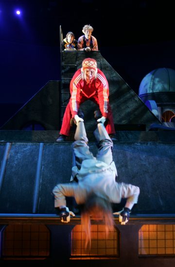 Neljä näyttelijää esiintyy dramaattisessa lavastuksessa; yksi punapukuinen vetää toista hahmoa roikkuen ylösalaisin katon reunan yli, kun taas kaksi muuta katselee ylemmältä tasanteelta, kaikki teatterivalaistuksessa.