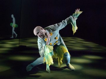 Teatteripukuinen esiintyjä, jolla on keltaisia ja vihreitä korostuksia, poseeraa dramaattisesti lavalla, ja lattialla on valonheittimen kuvio ja taustalla kävelee varjoinen hahmo.