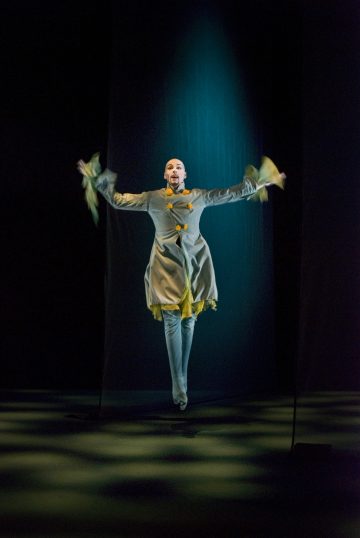 Teatteripukuinen esiintyjä, jolla on keltaisia korostuksia, hyppii kädet ojennettuina valonheittimen alla pimeällä näyttämöllä.