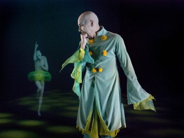 Etualalla seisoo mietteliäänä henkilö, jolla on vihreä puku ja keltaiset pompulat. Taustalla toinen vihreään tutuun pukeutunut hahmo poseeraa yhdellä jalalla ja nostetulla kädellä, molemmat tummaa näyttämöä vasten.