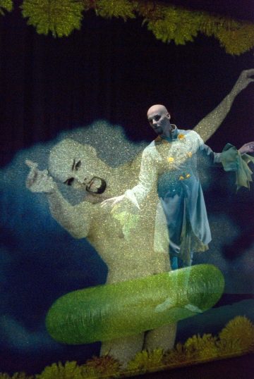 Valkoiseen pukuun ja dramaattiseen kasvomeikkiin pukeutunut esiintyjä poseeraa lavalla kädet ojennettuina. Hänen takanaan näkyy suuri, puoliksi läpinäkyvä alastonhahmon projektio ja vihreä esine, jotka luovat surrealistisen, taiteellisen kohtauksen.
