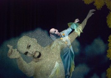 Siniseen pukuun ja kasvomaaliin pukeutunut esiintyjä poseeraa dramaattisesti lavalla, ja sen päällä on aavemainen, puoliksi läpinäkyvä kopio hänestä itsestään, mikä lisää kohtaukseen surrealistisen ja unenomaisen vaikutelman.