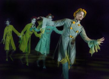 Pitkään, virtaavaan vihreään pukuun ja keltaiseen kukkakruunuun pukeutunut esiintyjä tanssii ojennetuin käsin pimeällä näyttämöllä jättäen jälkeensä värikkäitä aavemaisia jälkiä.