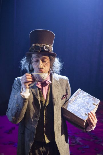 Steampunk-asuun pukeutunut mies, jolla on silinterihattu ja suojalasit, juo teekupista ja pitää kädessään vanhaa, kulunutta kirjaa. Hän seisoo dramaattisessa valaistuksessa punaisen ja violetin värisellä näyttämöllä.