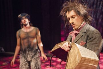 Pukuun pukeutunut epäsiisti mies lukee isosta, vanhasta kirjasta, kun taas toinen henkilö, jolla on karvaiset housut ja läpikuultava toppi, seisoo taustalla hämärästi valaistulla näyttämöllä.