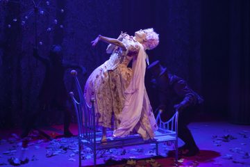 Nainen, jolla on koristeellinen historiallinen mekko ja peruukki, nojaa dramaattisesti taaksepäin sinisellä penkillä, jota ympäröi kaksi tummiin pukuihin pukeutunutta varjoista hahmoa lehdillä päällystetyllä ja violetilla valaistulla näyttämöllä.