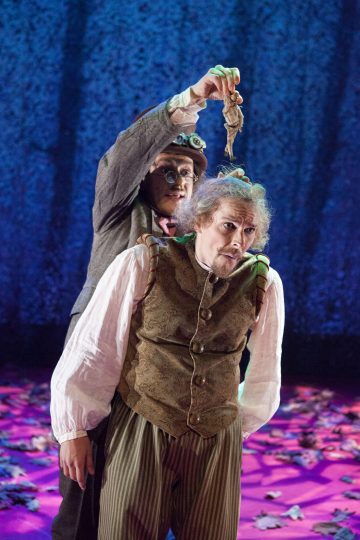 Kaksi näyttelijää näyttämöllä ajan puvuissa; toinen nostaa pienen nuken tai nuken toisen pään yläpuolelle. Taustalla on sinistä ja violettia, ja lattialle on ripoteltu lehtiä.