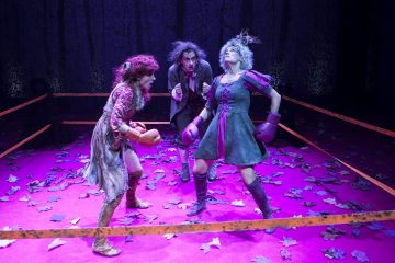 Kolme värikkäisiin pukuihin pukeutunutta näyttelijää seisoo nyrkkeilykehässä hajallaan olevien lehtien peittämällä näyttämöllä. Kahdella on nyrkkeilyhanskat ja he ovat vastakkain, kun taas kolmas seisoo heidän takanaan ja katsoo tarkkaavaisesti. Violetti valaistus määrittää näyttämön.