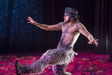 Vuohen näköiseen pukuun ja karvaisiin housuihin pukeutunut esiintyjä poseeraa näyttämöllä dramaattisesti, toinen jalka koholla ja kädet levällään, vaaleanpunaisessa valaistuksessa ja hajallaan olevien lehtien ympäröimänä.