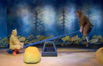 Kaksi eläinasuihin pukeutunutta ihmistä leikkii lavalla sinisellä keinulla, jonka lähellä on maalattu metsätausta ja keltaisia kiviä muistuttavia rekvisiittaa. Valaistus luo omituisen tunnelman.