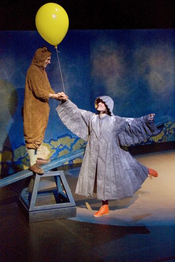 Karhupukuinen henkilö, jolla on keltainen ilmapallo kädessään, seisoo keinun toisessa päässä, kun taas toinen henkilö, jolla on harmaa lintupuku ja oranssit kengät, tasapainoilee toisessa päässä maalatun teatteritaustan edessä.