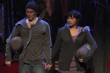 Mies ja nainen seisovat kädestä pitäen, kummallakin on rullattu makuupussi. Miehellä on pipo ja harmaa takki, naisella turkisvuorillinen hupparipusero. He näyttävät vakavilta ja katsovat eteenpäin hämärässä valaistussa ympäristössä.