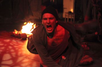 Pipoon ja avoimeen takkiin pukeutunut mies huutaa intohimoisesti eteenpäin nojautuen suu auki. Taustalla palaa tuli, joka heittää dramaattisen punaisen hehkun koko kohtaukseen.