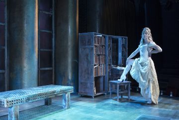 Vanhanaikaiseen, hienostuneeseen mekkoon pukeutunut nainen poseeraa toinen jalka tuolilla avoimen kirjakaapin vieressä hämärästi valaistulla näyttämöllä, jossa on suuret pylväät ja ruudullinen penkki.