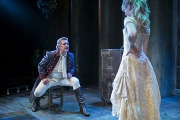 Aikakauden pukuihin pukeutunut mies istuu jakkaralla, nojaa eteenpäin ja katsoo keskittyneesti valkoiseen mekkoon pukeutunutta naista, joka seisoo kädet lanteillaan hämärästi valaistulla näyttämöllä, jonka takana on suuri arkku.