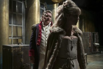 Korsettimekkoon ja turkisverhoiltuun viittaan pukeutuneen naisen takana seisoo koristeltuun sotilasunivormuun pukeutunut mies dramaattisessa, hämärässä lavastuksessa, jossa on kultaisia pylväitä ja arkunpäätä. Nainen katsoo poispäin, näyttää järkyttyneeltä tai etäiseltä.