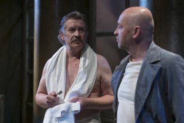 Kaksi miestä seisoo sisätiloissa; toinen on ilman paitaa, pyyhe hartioillaan, ja hänellä on partaveitsi kädessään, kun taas toinen, kalju ja harmaaseen takkiin pukeutunut mies on häntä vastassa. Molemmat vaikuttavat vakavilta, kuin olisivat keskustelemassa.