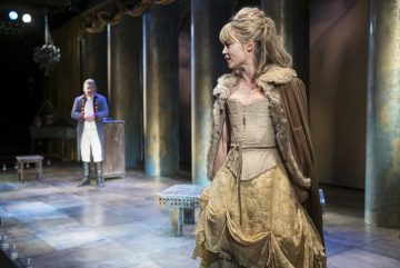 Kultaiseen aikakauden pukuun ja viittaan pukeutunut nainen seisoo lavalla sivuttain, ja taustalla seisoo historialliseen sotilaspukuun pukeutunut mies. Lavastuksessa on korkeat pylväät ja dramaattinen valaistus, joka tuo mieleen historiallisen tai klassisen näytelmän.