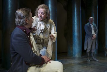 Kaksi aikakauden pukuihin pukeutunutta näyttelijää käyvät näyttämöllä kiihkeää keskustelua, toinen istuu ja kuuntelee, kun toinen elehtii vilkkaasti. Kolmas näyttelijä seisoo taustalla, osittain varjossa, ja tarkkailee kohtausta.