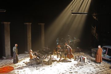 Dramaattinen näyttämökohtaus, jossa on viisi esiintyjää: pianisti, kaksi jousimuusikkoa ja kaksi näyttelijää. Valonsäteet valaisevat heitä, ja taustalla on pylväitä ja vaurioitunut auto, jotka luovat tunnelmallisen ilmapiirin.