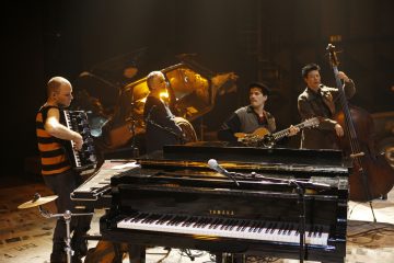 Neljä muusikkoa esiintyy lavalla eri soittimilla, muun muassa harmonikalla, sähkökitaralla, pystybassolla ja etualalla olevalla Yamaha-pianolla. Taustalla on osittain murskattu auto ja hämärä, dramaattinen valaistus.