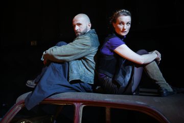 Mies ja nainen istuvat selkä vastakkain ruosteisen auton katolla ja katsovat vastakkaisiin suuntiin. Miehellä on vihreä takki ja ajeltu pää, kun taas naisella on musta liivi ja ruskeat saappaat. Tausta on tumma.