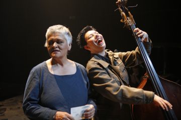 Kaksi ihmistä lavalla: toisella on lyhyet harmaat hiukset ja sininen toppi, kädessä paperi, ja toisella on lyhyet tummat hiukset, takki päällä, hymyilee leveästi ja soittaa kontrabassoa. Heidät valaistaan näyttämövaloilla.