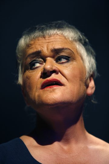 Henkilö, jolla on lyhyet harmaat hiukset, dramaattinen meikki ja tumma yläosa, katsoo ylöspäin vakavalla ilmeellä tummaa taustaa vasten.