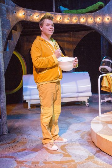 Keltaiseen verryttelypukuun pukeutunut henkilö seisoo värikkäällä näyttämöllä, kädessään valkoinen kulho ja lusikka, kirkkaassa valaistuksessa ja taustalla on futuristisia lavaste-elementtejä.