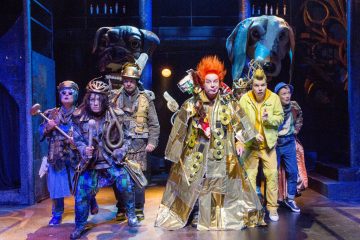 Lavalla seisoo kuusi näyttelijää värikkäissä, omalaatuisissa puvuissa, ja joillakin on kädessään rekvisiittaa. Taustalla on kaksi suurta koiranpääveistosta, jotka luovat dramaattisen, omituisen teatterikohtauksen eloisalla valaistuksella.