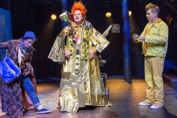 Lavalla esiintyy kolme näyttelijää. Keskimmäisellä on kiiltävä kultatakki ja villi oranssi peruukki, ja häntä reunustavat kaksi muuta näyttelijää, joilla on rennot puvut; toinen kantaa sinistä laukkua ja toinen vihreää esinettä. Kohtaus on kirkkaasti valaistu.