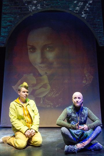 Kaksi ihmistä istuu ristissä lavalla naisen kasvojen heijastaman kuvan alla. Toisella on keltainen asu, huppari ja lyhyet hiukset, toisella on kuvioitu sinivihreä asu ja hän on kalju. Tausta on tumma ja teatraalinen.