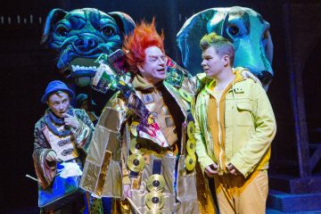 Lavalla esiintyy värikkäisiin pukuihin pukeutuneita näyttelijöitä; yhdellä on kirkkaanpunainen tukka ja metallinen viitta, jossa on CD-levyjä, toisella on keltainen, ja taustalla on kaksi suurta koiranukkea ja muusikko huilun kanssa.