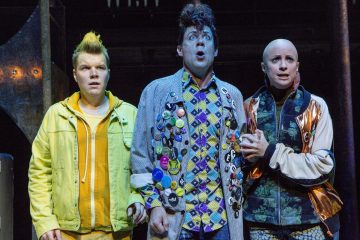 Kolme näyttelijää värikkäissä ja omalaatuisissa puvuissa reagoi näyttämöllä yllättyneenä ja huolestuneena. Vasemmalla olevalla henkilöllä on keltainen piikkitukka, keskellä olevalla on kihara tukka ja nappien peittämä takki, ja oikealla on kalju, joka puristaa keskellä olevan henkilön kättä.