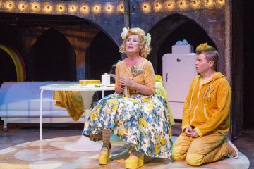 Värikkääseen mekkoon ja keltaisiin kenkiin pukeutunut nainen istuu lavalla keltaiseen huppariin pukeutuneen nuoren vieressä, ja heidän takanaan on valkoinen jääkaappi ja pöytä lämpimässä valaistuksessa.