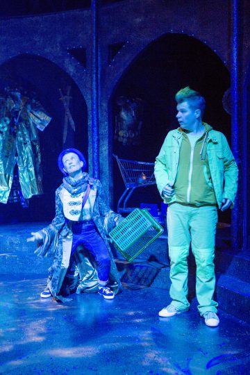 Kaksi ihmistä seisoo sinisen näyttämövalaistuksen alla. Toisella on pitkä kuviollinen takki ja hattu, ja hän pitelee koria dramaattisesti. Toinen, vihreään pukeutunut ja juoma kädessään, katsoo häntä hämmästyneenä. Taustalla on kiiltäviä pukuja ja ostoskärry.