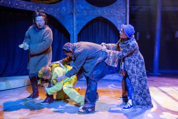 Lavalla esiintyy neljä näyttelijää värikkäissä, omituisissa puvuissa. Yksi keltaisiin pukeutunut näyttelijä on kyyristynyt, kun kaksi muuta vetää vaatteistaan, ja neljäs seisoo lähellä, ja kaikilla on ilmeikkäät kasvot ja dynaamiset asennot näyttämövalaistuksessa.