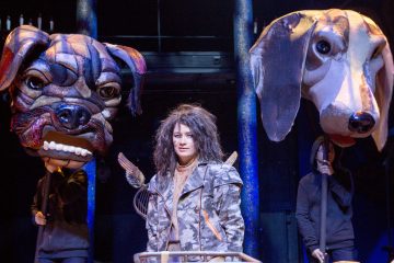 Naamiotakkiin pukeutunut henkilö seisoo lavalla kahden jättimäisen koiranpäänuken välissä, joita kumpaakin ohjaa mustiin pukeutunut henkilö, tummaa teatteritaustaa vasten.