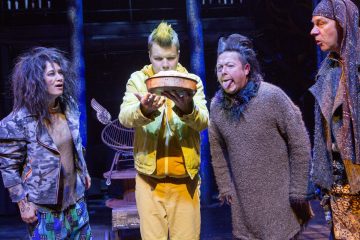 Lavalla seisoo neljä näyttelijää värikkäissä puvuissa ja peruukissa. Yksi keltapukuinen pitää piirakkaa kädessään, ja toinen harmaapukuinen työntää kielensä ulos, ja kaikki vaikuttavat uteliailta tai innokkailta. Näyttämön taustalla on maalaismaista rekvisiittaa ja sininen valaistus.