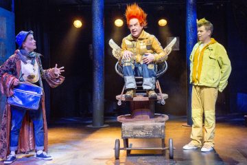 Lavalla esiintyy kolme näyttelijää. Yhdellä on värikäs takki ja sininen laukku, toinen istuu siipituolissa kirkkaan oranssi piikkitukka, ja kolmannella on keltaiset vaatteet ja vihreät hiukset. Dramaattinen valaistus korostaa heitä.