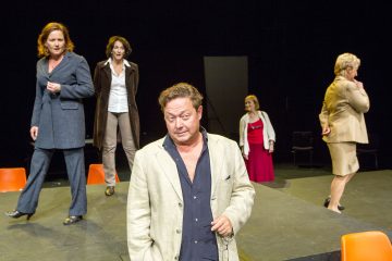 Viisi näyttelijää seisoo oransseilla tuoleilla varustetulla näyttämöllä liike- ja juhla-asuihin pukeutuneina ja näyttävät esittävän intensiivisen tai dramaattisen kohtauksen. Edessä olevalla miehellä on beige takki ja hän katsoo alaspäin, kun taas muut ovat hänen takanaan välissä.