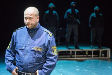 Etualalla seisoo siniseen poliisin univormuun pukeutunut mies, joka katsoo sivulle. Kolme tummiin hupparivaatteisiin pukeutunutta ihmistä seisoo varjoisassa taustalla. Kohtaus on hämärästi valaistu ja näyttää olevan näyttämöllä.