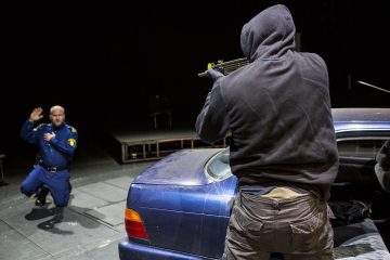 Huppariin pukeutunut henkilö tähtää aseella siniseen univormuun pukeutunutta polvistuvaa miestä, jolla on kädet kohotettuna sinisen auton vieressä hämärässä näyttämöllä.